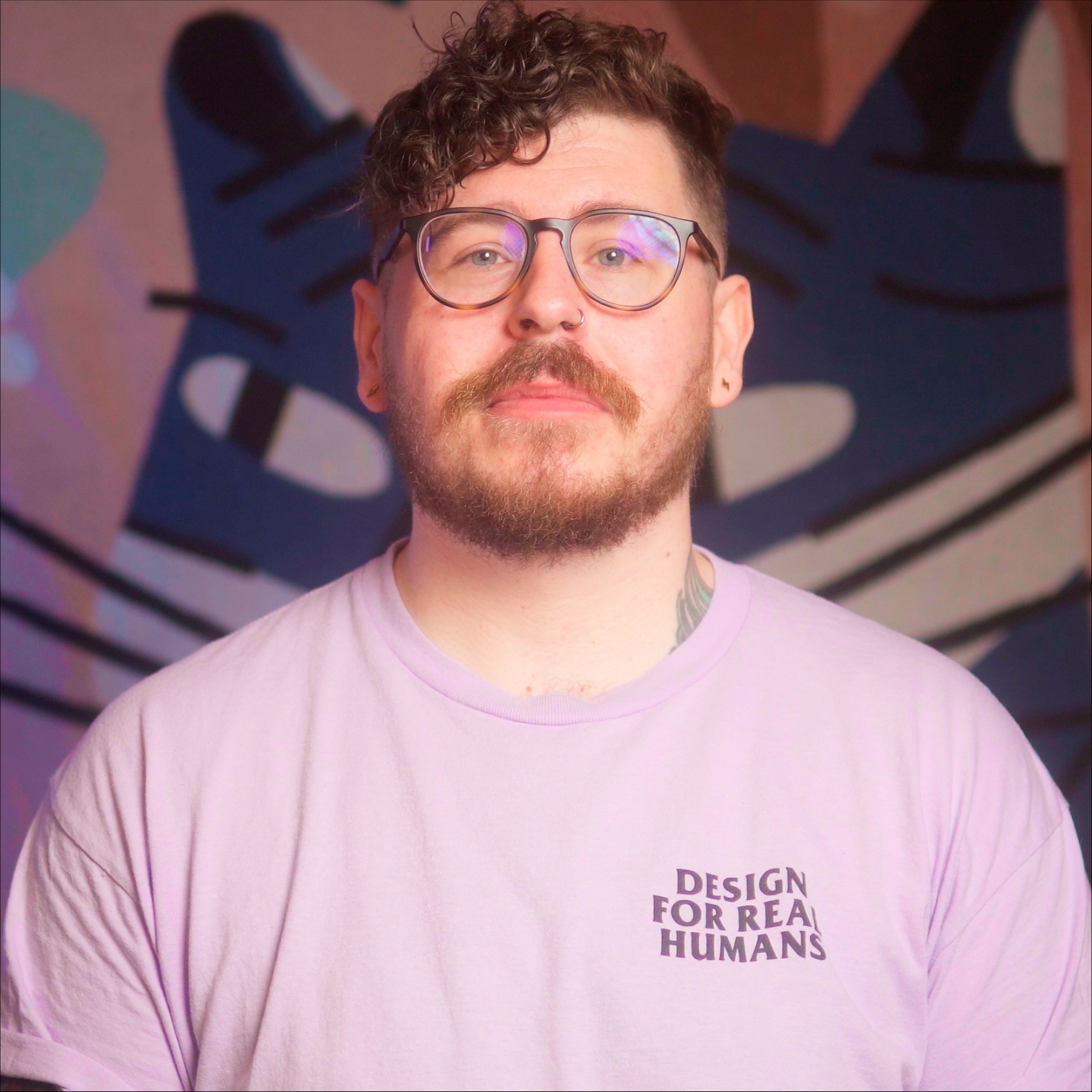 Profile shot of Scott Riley. Curly-haired lad wearing glasses and a purple "design for real humans" t-shirt, poses earnestly for his school photograph.